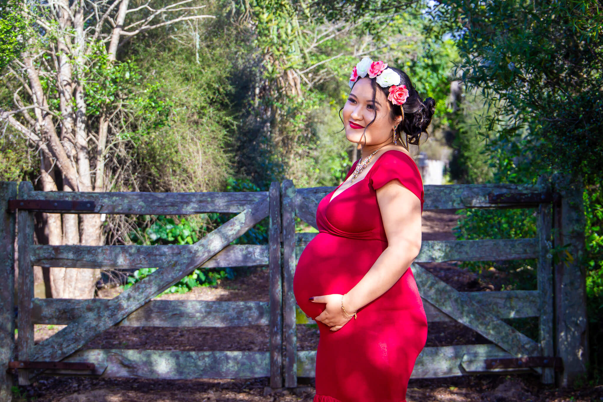 Auckland maternity photographer — woman in red gown with floral crown at rustic gate